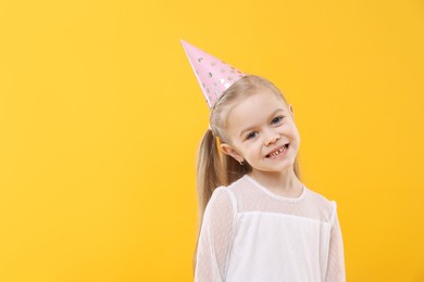 Photo of Cute little girl in conical paper hat on yellow background, space for text. Birthday surprise party