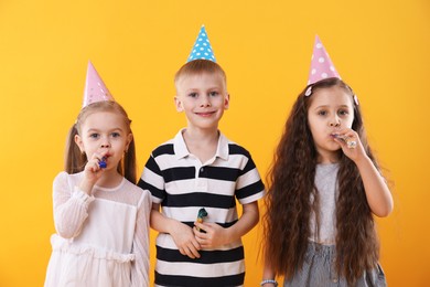 Photo of Children in conical paper hats with blowers on yellow background. Birthday surprise party