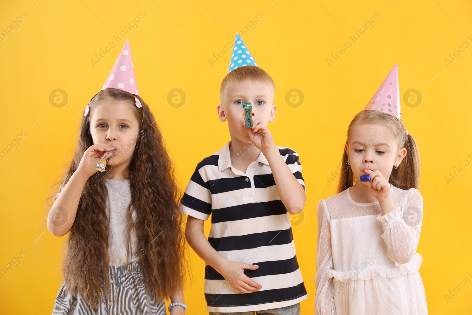 Photo of Children in conical paper hats with blowers on yellow background. Birthday surprise party