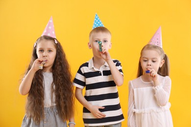 Photo of Children in conical paper hats with blowers on yellow background. Birthday surprise party