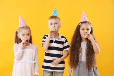 Photo of Children in conical paper hats with blowers on yellow background. Birthday surprise party