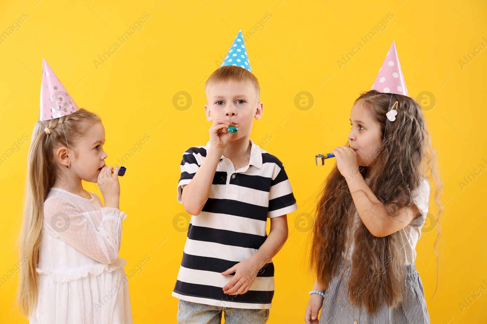 Photo of Children in conical paper hats with blowers on yellow background. Birthday surprise party