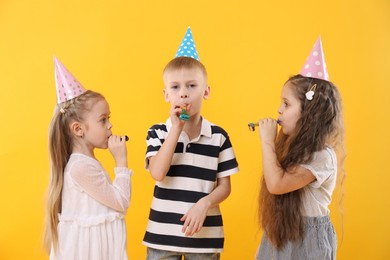 Photo of Children in conical paper hats with blowers on yellow background. Birthday surprise party