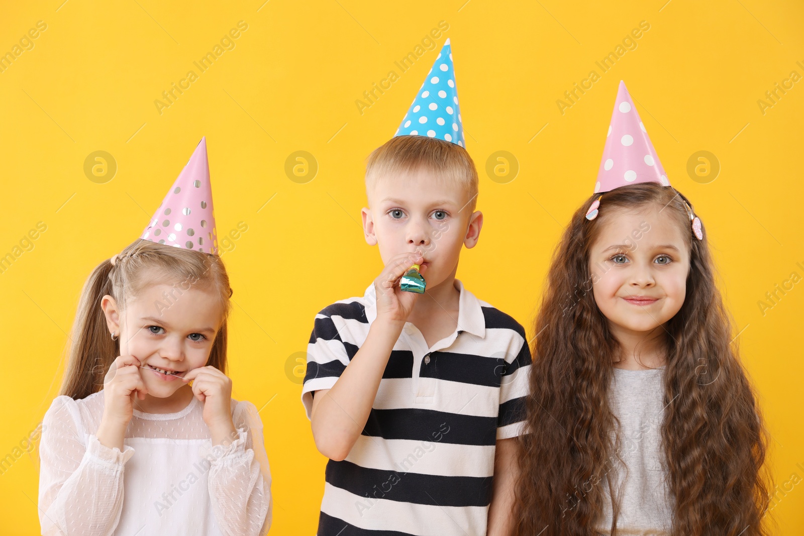 Photo of Children in conical paper hats with blower on yellow background. Birthday surprise party