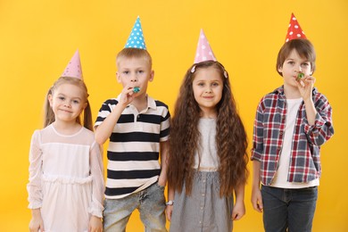 Photo of Children in conical paper hats with blowers on yellow background. Birthday surprise party