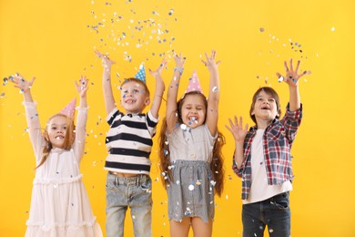 Photo of Happy children in conical paper hats under falling confetti on yellow background. Birthday surprise party