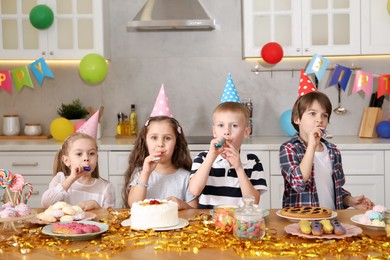 Photo of Children in conical paper hats with blowers celebrating birthday indoors. Surprise party