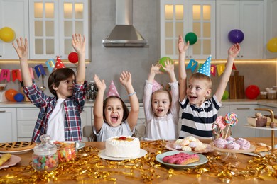 Photo of Cheerful children at table with tasty cake and different treats celebrating birthday indoors. Surprise party