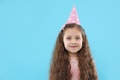 Photo of Cute little girl in conical paper hat on light blue background, space for text. Birthday surprise party