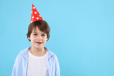 Photo of Cute little boy in conical paper hat on light blue background, space for text. Birthday surprise party