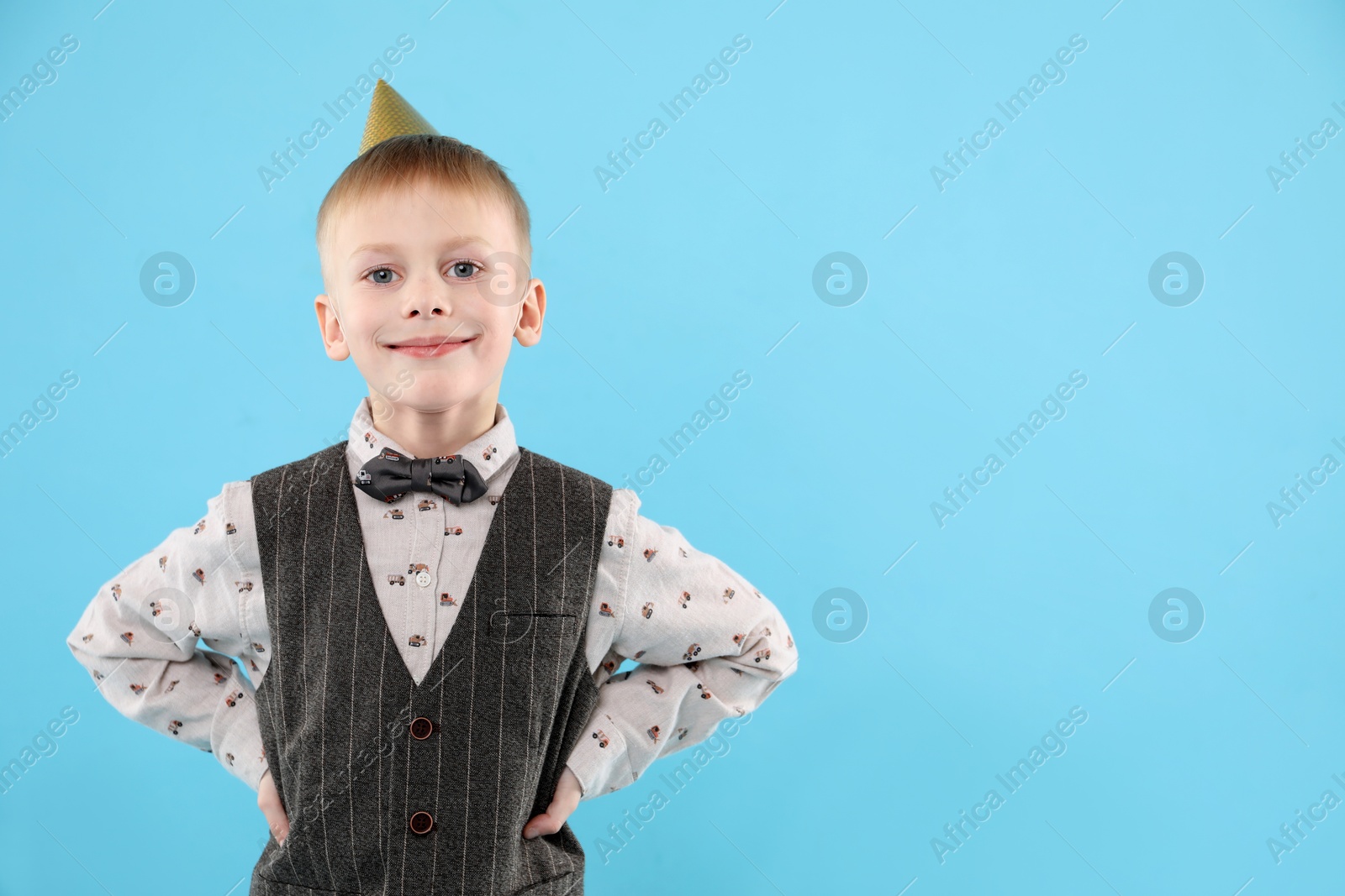 Photo of Cute little boy in conical paper hat on light blue background, space for text. Birthday surprise party