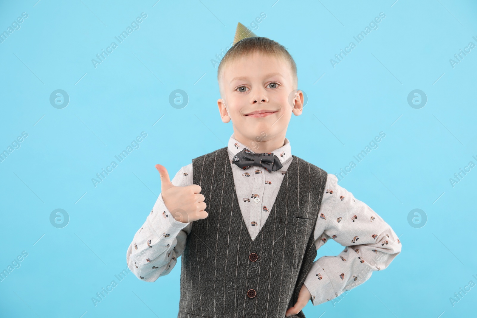 Photo of Cute little boy in conical paper hat showing thumbs up on light blue background. Birthday surprise party