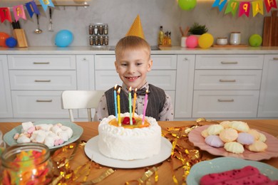 Photo of Cute little boy at table with birthday cake and different treats indoors. Surprise party