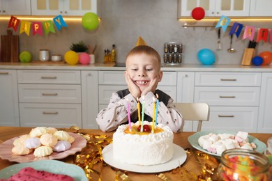 Photo of Cute little boy at table with birthday cake and different treats indoors. Surprise party