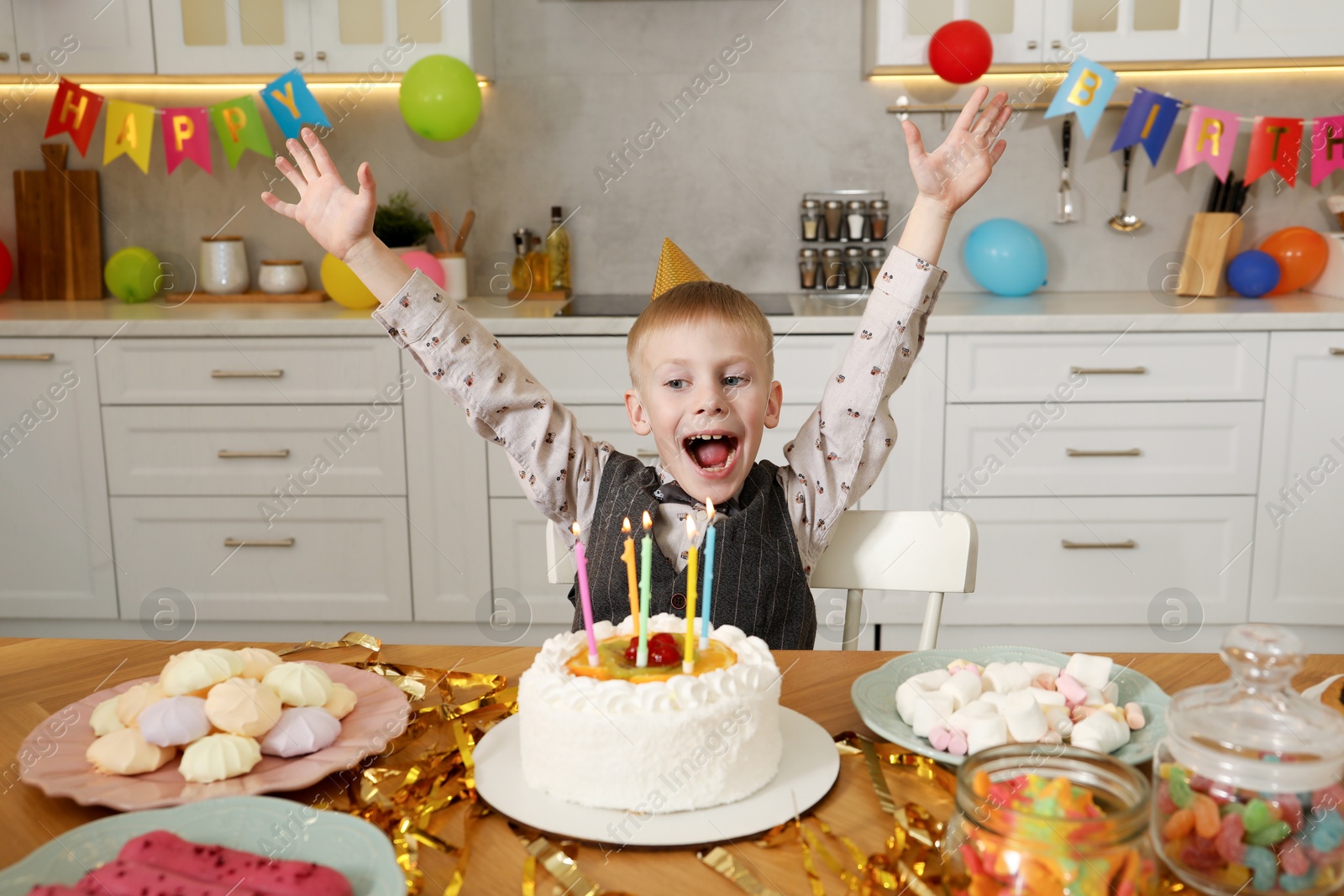 Photo of Cute little boy at table with birthday cake and different treats indoors. Surprise party