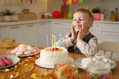 Photo of Cute little boy at table with birthday cake and different treats indoors. Surprise party