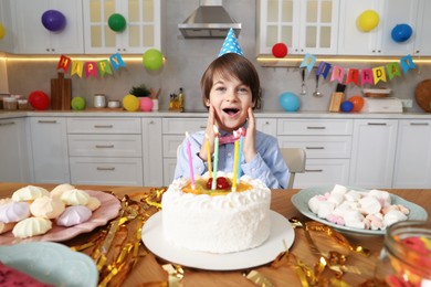 Photo of Cute little boy at table with birthday cake and different treats indoors. Surprise party