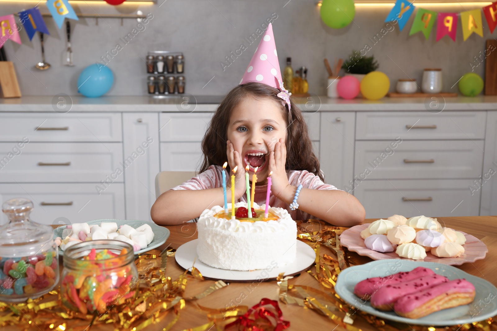 Photo of Cute little girl at table with birthday cake and different treats indoors. Surprise party