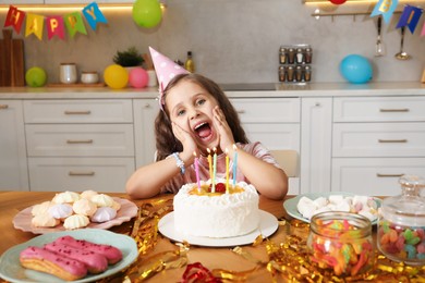 Photo of Cute little girl at table with birthday cake and different treats indoors. Surprise party