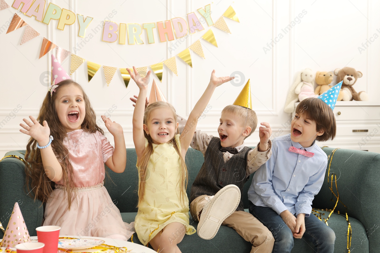 Photo of Cheerful children in conical paper hats celebrating birthday indoors. Surprise party