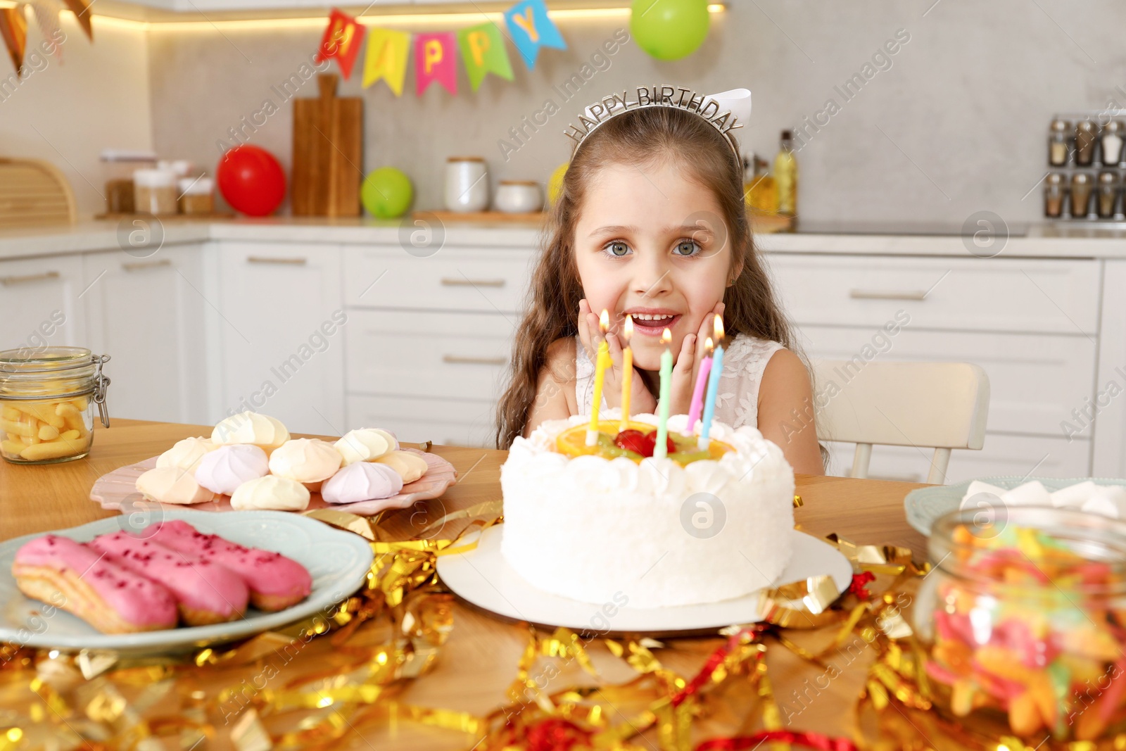 Photo of Cute little girl at table with birthday cake and different treats indoors. Surprise party