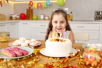 Photo of Cute little girl at table with birthday cake and different treats indoors. Surprise party