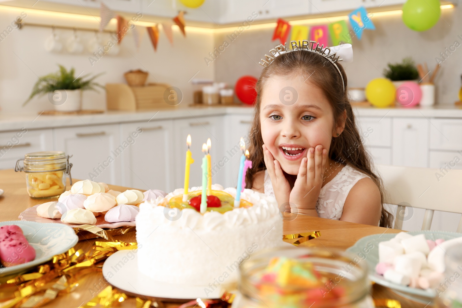 Photo of Cute little girl at table with birthday cake and different treats indoors. Surprise party