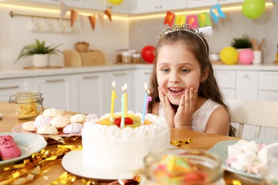 Photo of Cute little girl at table with birthday cake and different treats indoors. Surprise party