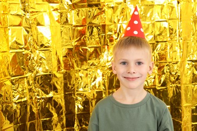 Photo of Cute little boy in conical paper hat near golden foil curtain, space for text. Birthday surprise party