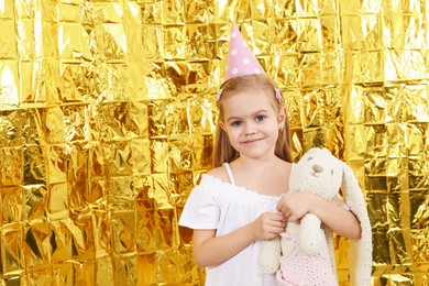 Photo of Cute little girl in conical paper hat with toy bunny near golden foil curtain, space for text. Birthday surprise party