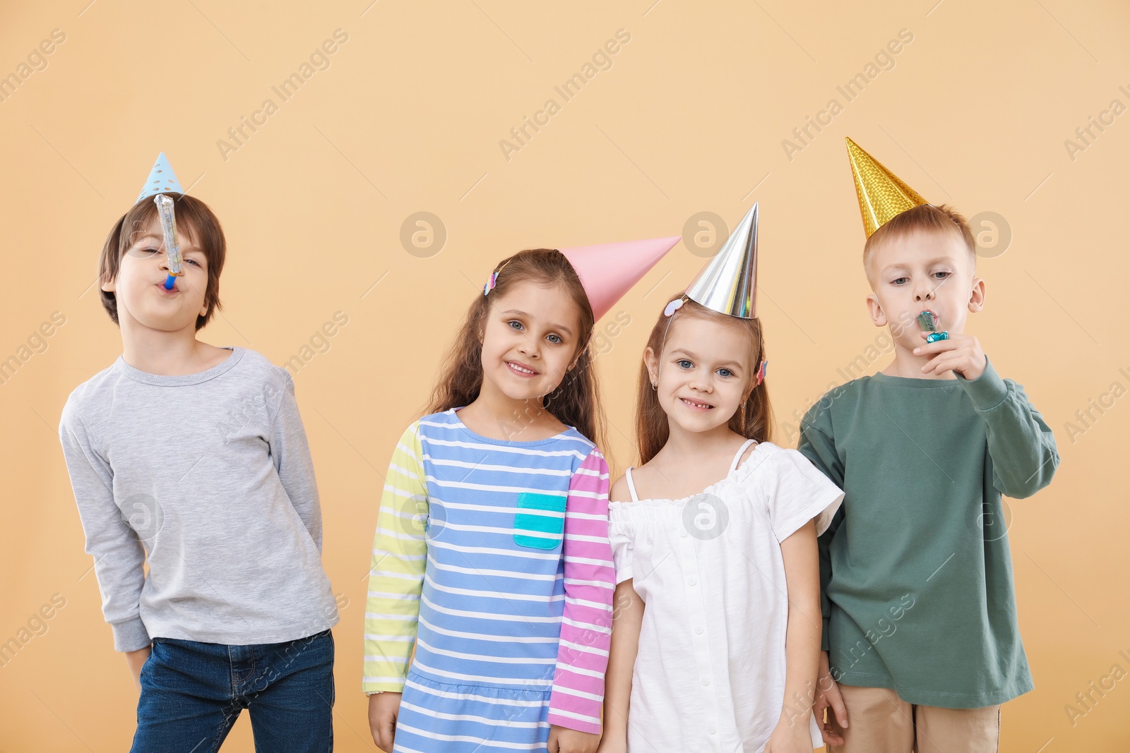 Photo of Children in conical paper hats with blowers on beige background. Birthday surprise party