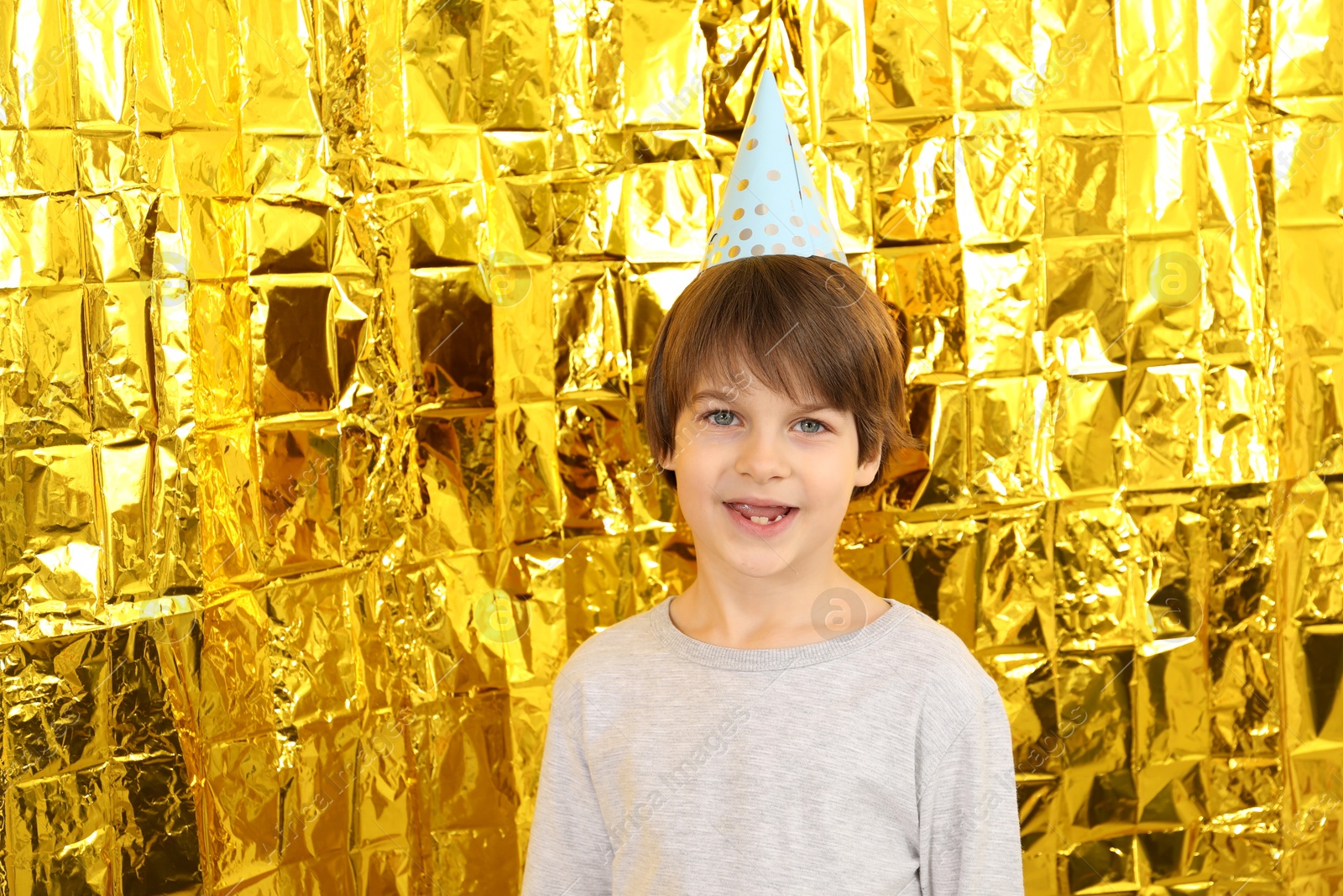 Photo of Cute little boy in conical paper hat near golden foil curtain. Birthday surprise party