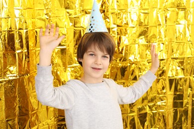 Photo of Cute little boy in conical paper hat near golden foil curtain. Birthday surprise party