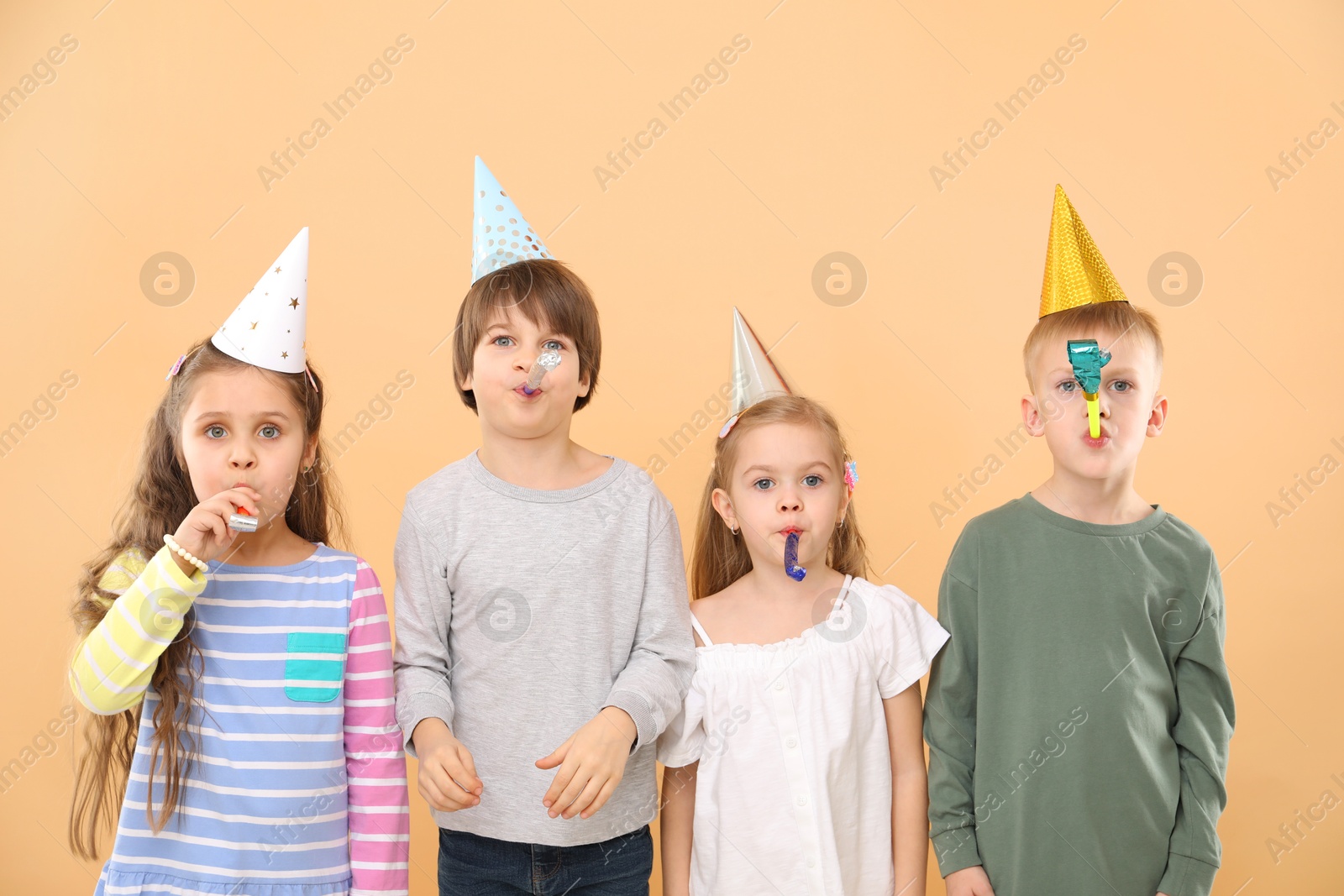 Photo of Children in conical paper hats with blowers on beige background. Birthday surprise party