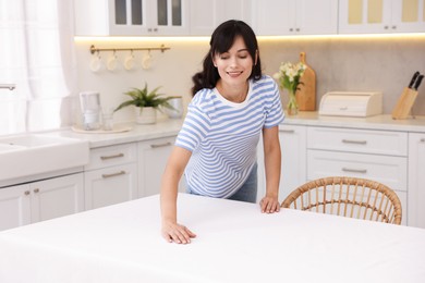 Photo of Young woman putting white tablecloth on table in kitchen