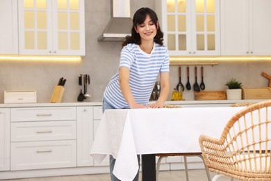 Photo of Young woman putting white tablecloth on table in kitchen