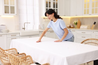 Photo of Young woman putting white tablecloth on table in kitchen