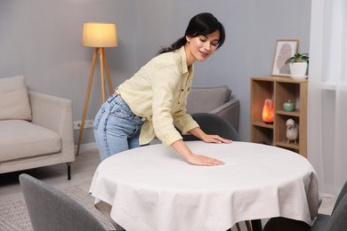 Photo of Young woman putting white tablecloth on table at home