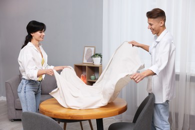 Photo of Couple putting white tablecloth on table at home