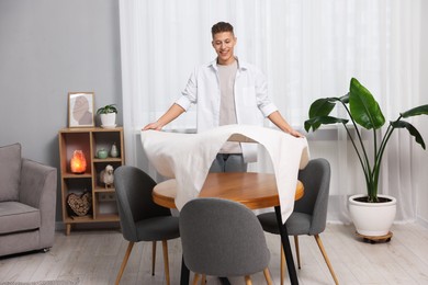 Photo of Young man putting white tablecloth on table at home