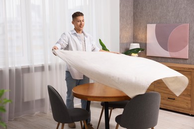 Photo of Young man putting white tablecloth on table at home