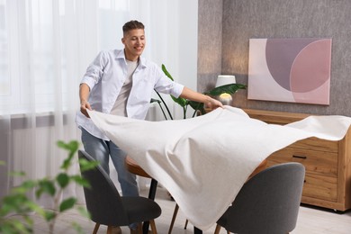 Photo of Young man putting white tablecloth on table at home