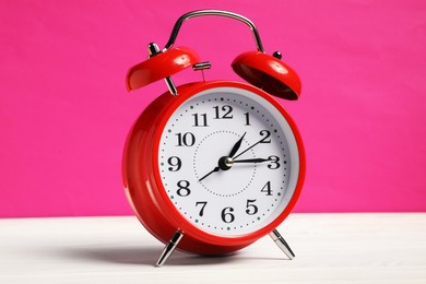 Photo of One red alarm clock on white wooden table against bright pink background