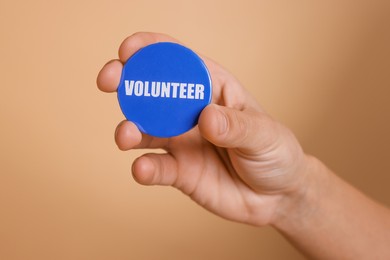 Photo of Man holding button badge with word Volunteer on beige background, closeup