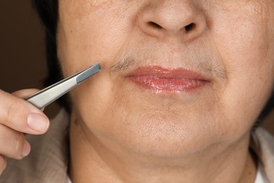 Photo of Senior woman plucking her mustache with tweezers on brown background, closeup