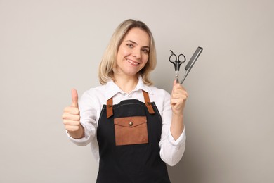 Photo of Smiling hairdresser with comb and scissors showing thumbs up on gray background