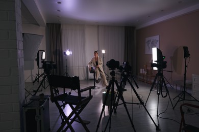 Photo of Young woman sitting on chair near cameras and other professional equipment in modern film studio