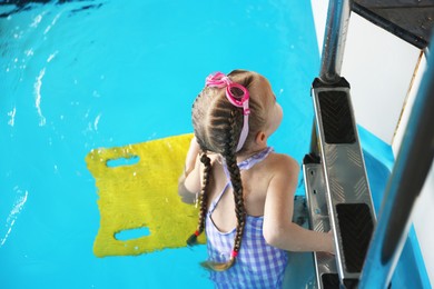 Photo of Little girl with goggles and kickboard swimming in pool indoors, above view