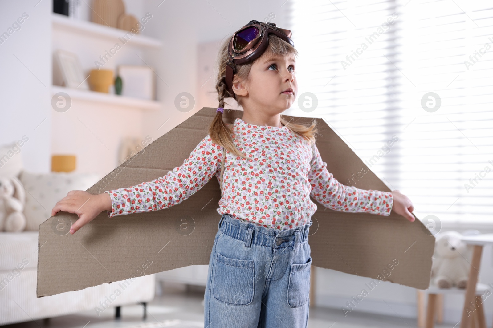 Photo of Cute little girl with cardboard plane wings and goggles playing pilot at home