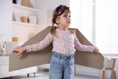 Photo of Cute little girl with cardboard plane wings and goggles playing pilot at home